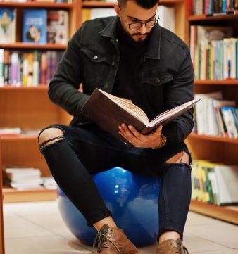 Tall smart arab student man, wear on black jeans jacket and eyeglasses, sitting at library and read book at hands.
